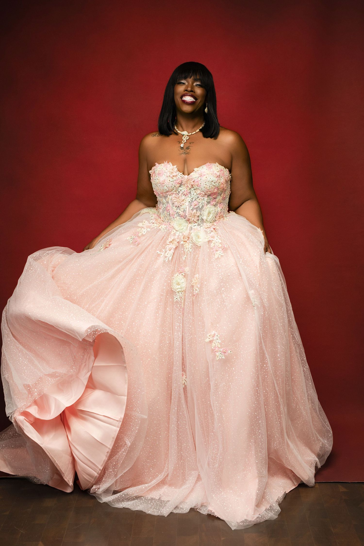 A plus-size woman in a strapless pink floral ball gown poses confidently against a red background.