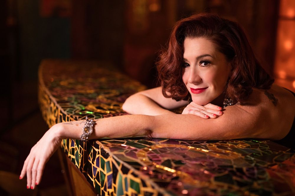 A smiling woman with red hair leans on a colorful mosaic bar counter in a warmly lit venue.