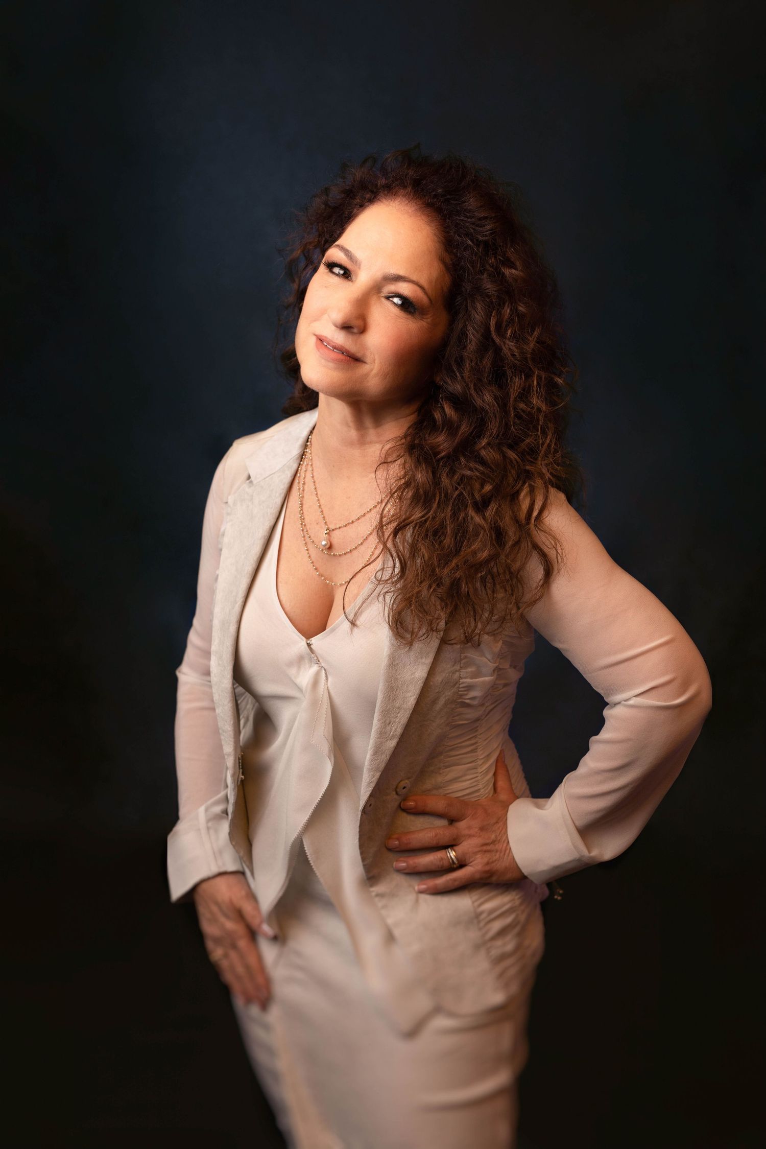 Woman with curly hair poses confidently in white blazer and dress against dark studio background.
