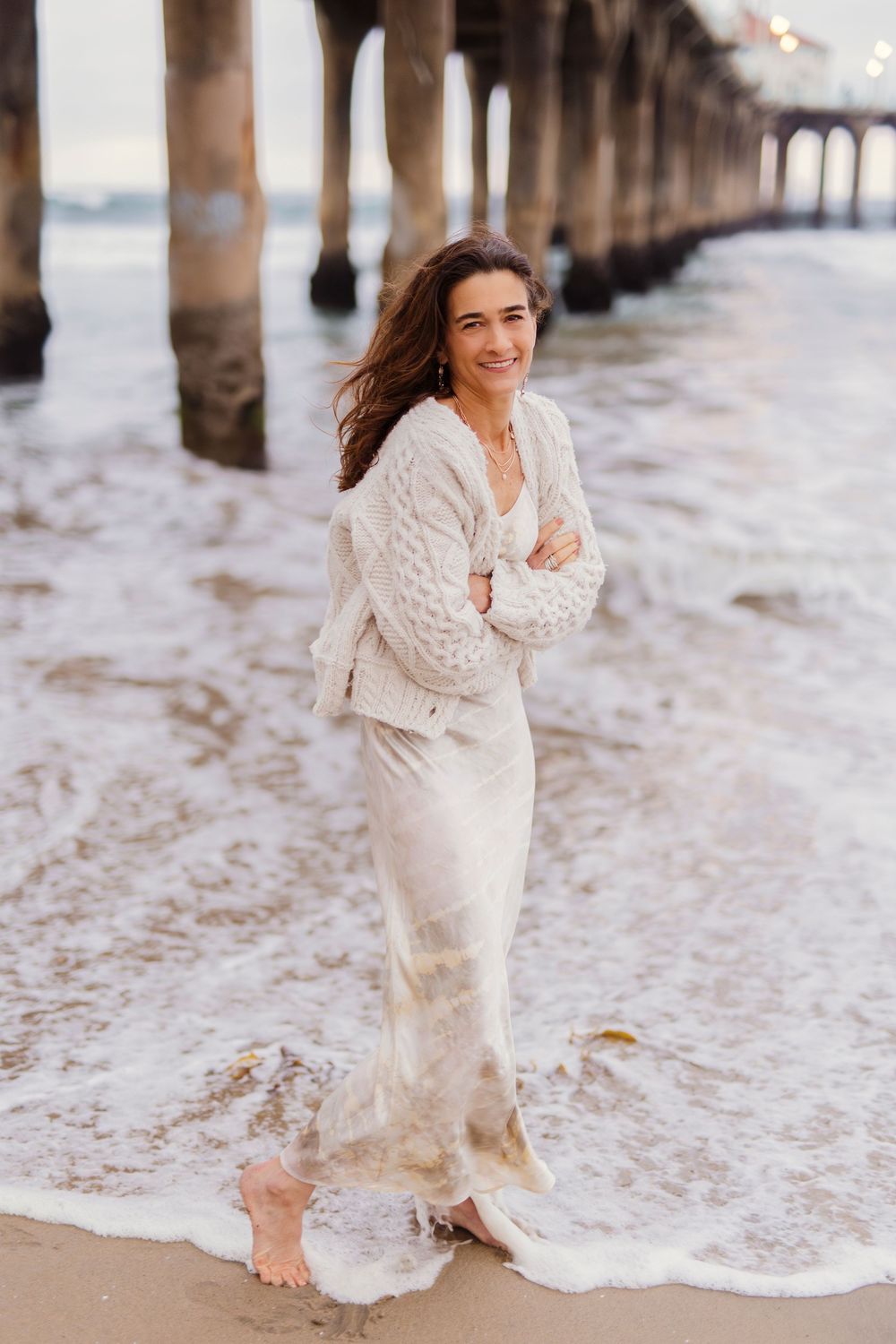 Person wearing cozy white knit sweater poses on beach at sunset with pier columns and ocean waves in background.