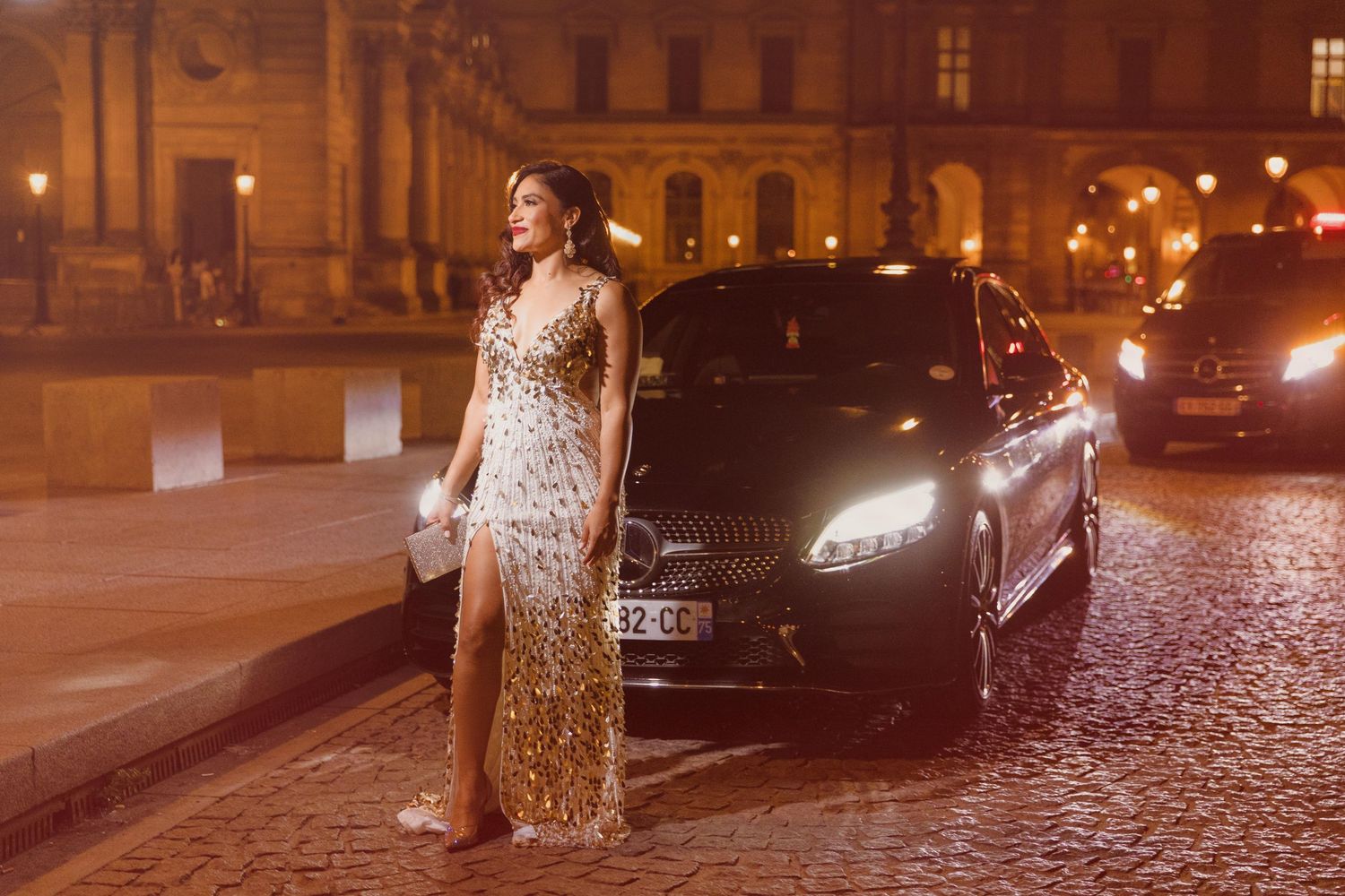 Woman in sequined gown poses beside luxury car on cobblestone street at night near illuminated building.