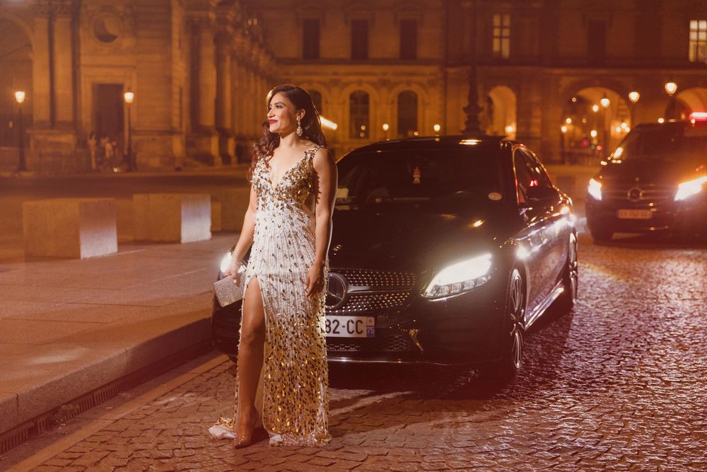 Woman in sequined gown poses beside luxury car on cobblestone street at night near illuminated building.