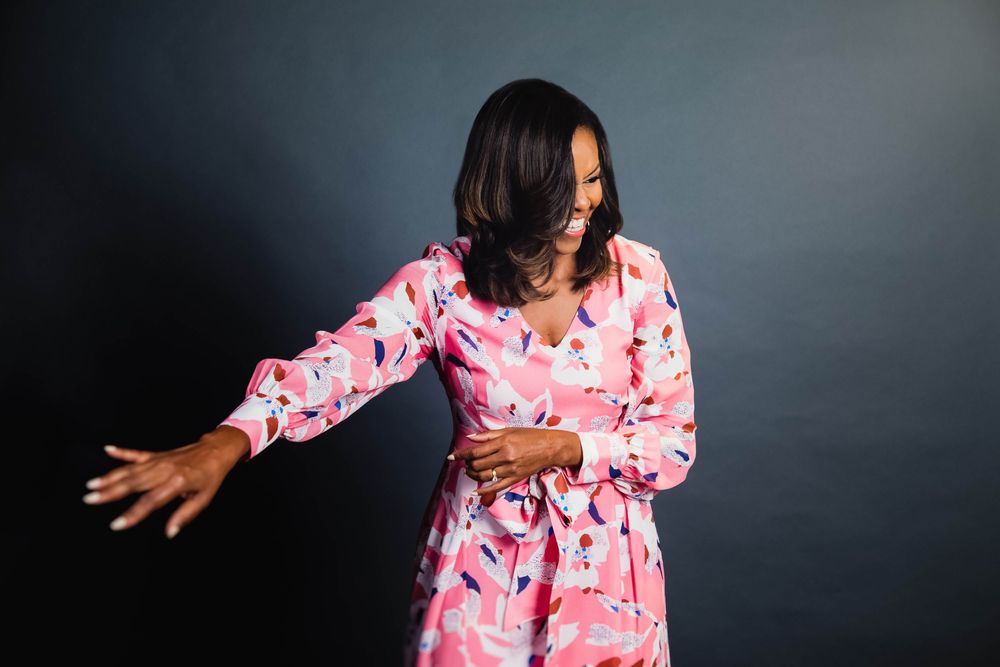 Woman in pink floral dress posing gracefully against dark background, extending arm outward with a joyful smile.
