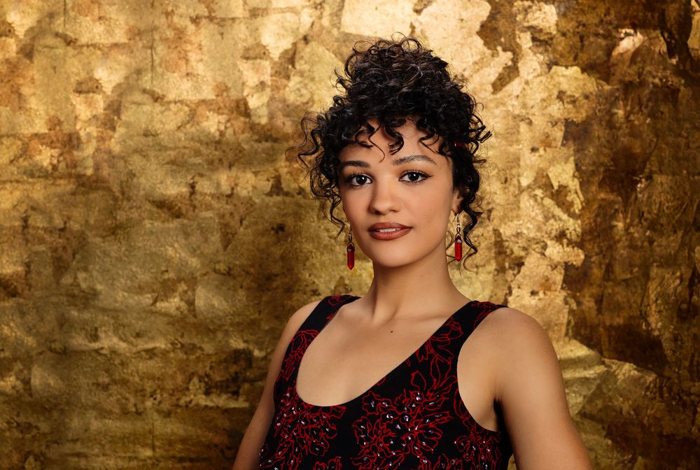 Person wearing a red sequin dress poses elegantly against a textured gold wall in dramatic lighting.