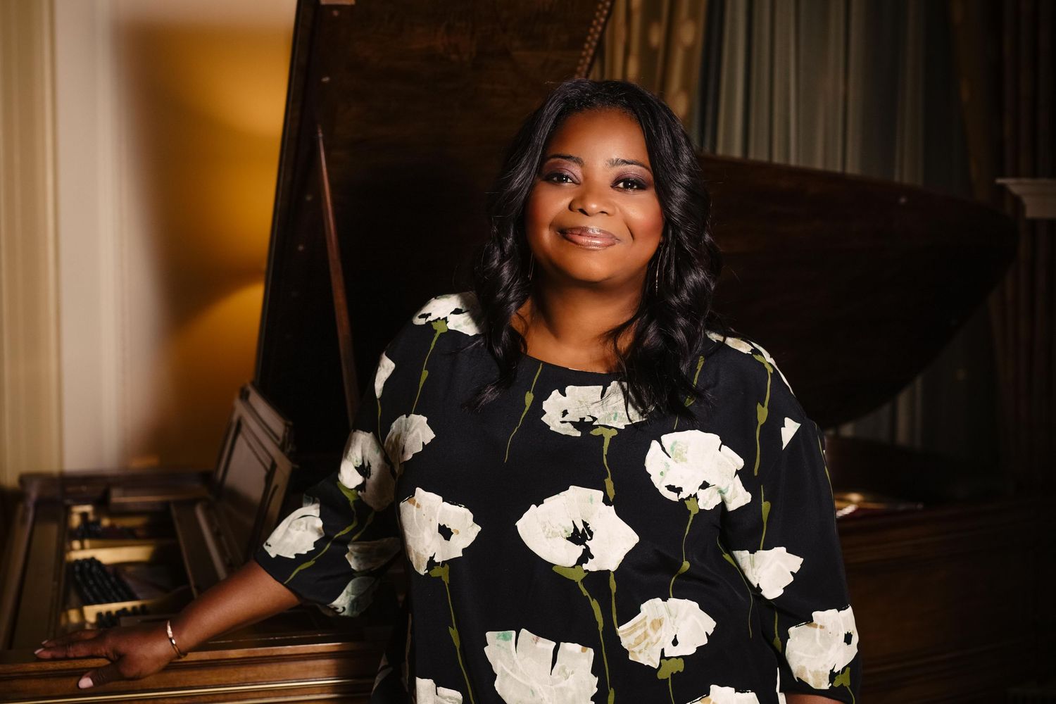 Woman in black floral dress smiling beside a grand piano in an elegant room.