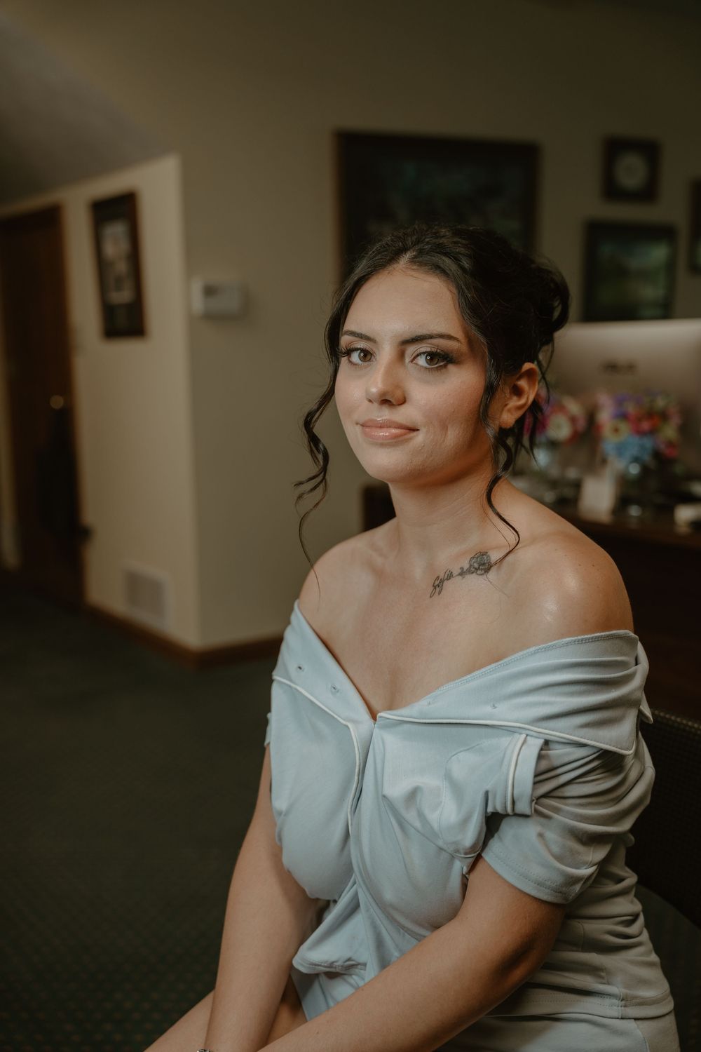 Elegant bridesmaid in off-shoulder silver dress with neck tattoo poses gracefully in an indoor setting.