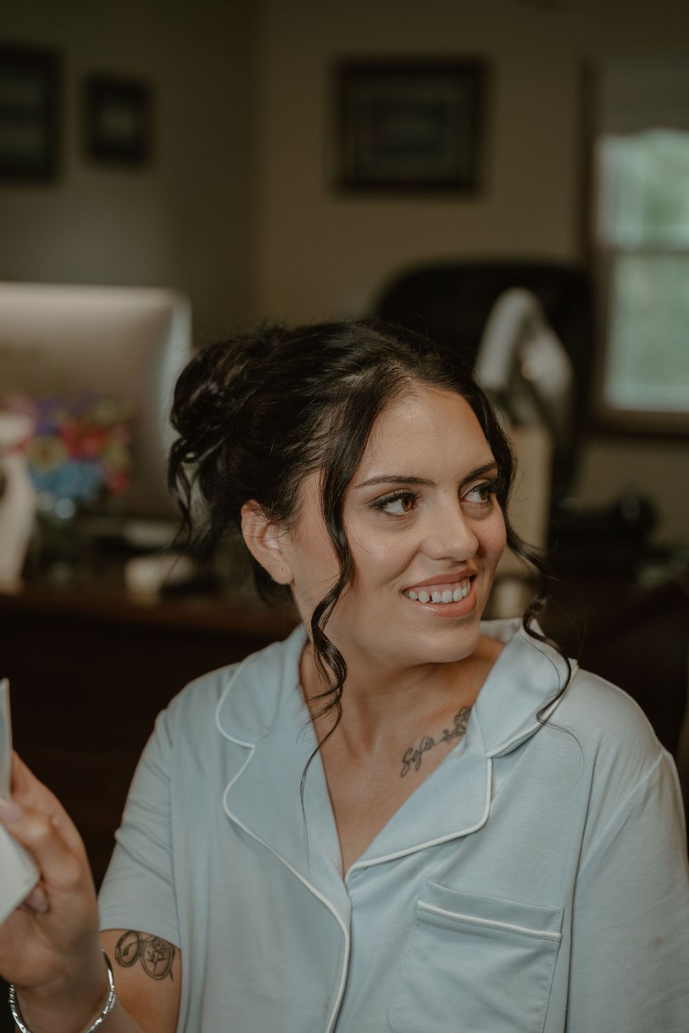 Smiling bridesmaid with dark updo hair and neck tattoo wearing white pajamas reads a card indoors.
