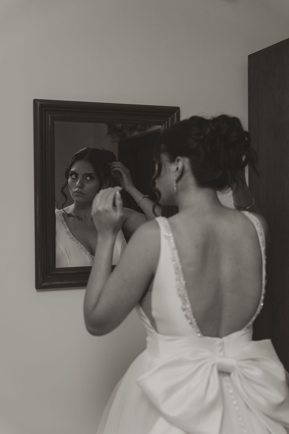 Bride in backless wedding gown adjusts her hair while gazing into a framed mirror, captured in black and white.