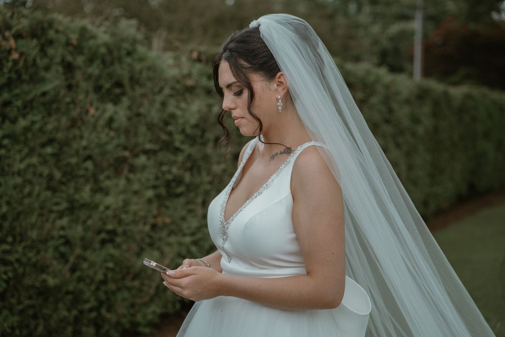 Elegant bride in a white gown and long veil looks down at her phone in a lush garden setting.