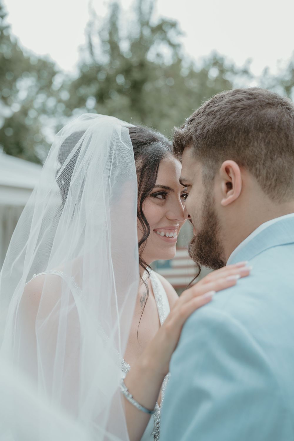Smiling bride and bearded groom share an intimate moment under a sheer wedding veil outdoors.