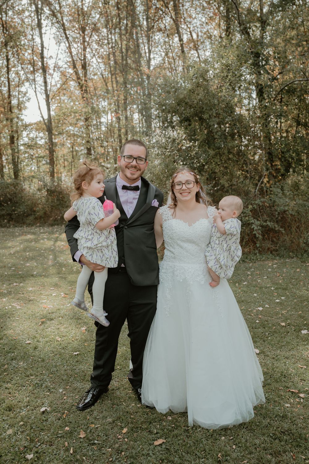 Bride and groom in wedding attire smile outdoors holding two young children in an autumn woodland setting.