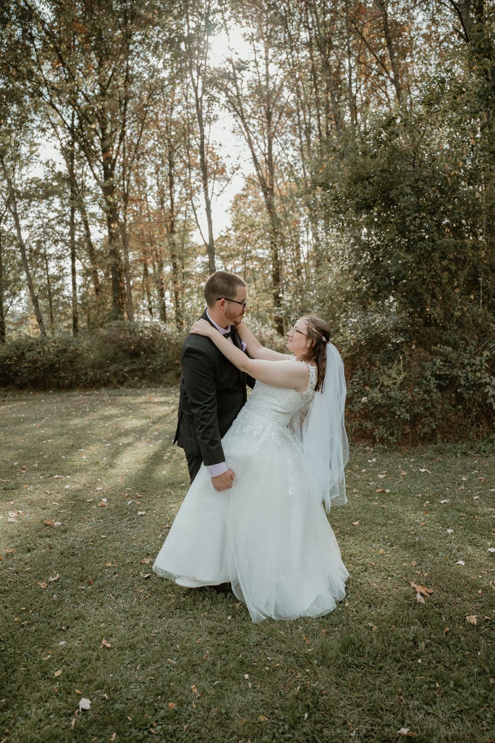 Bride in white lace gown and groom in black tuxedo dance together on grass surrounded by autumn trees.