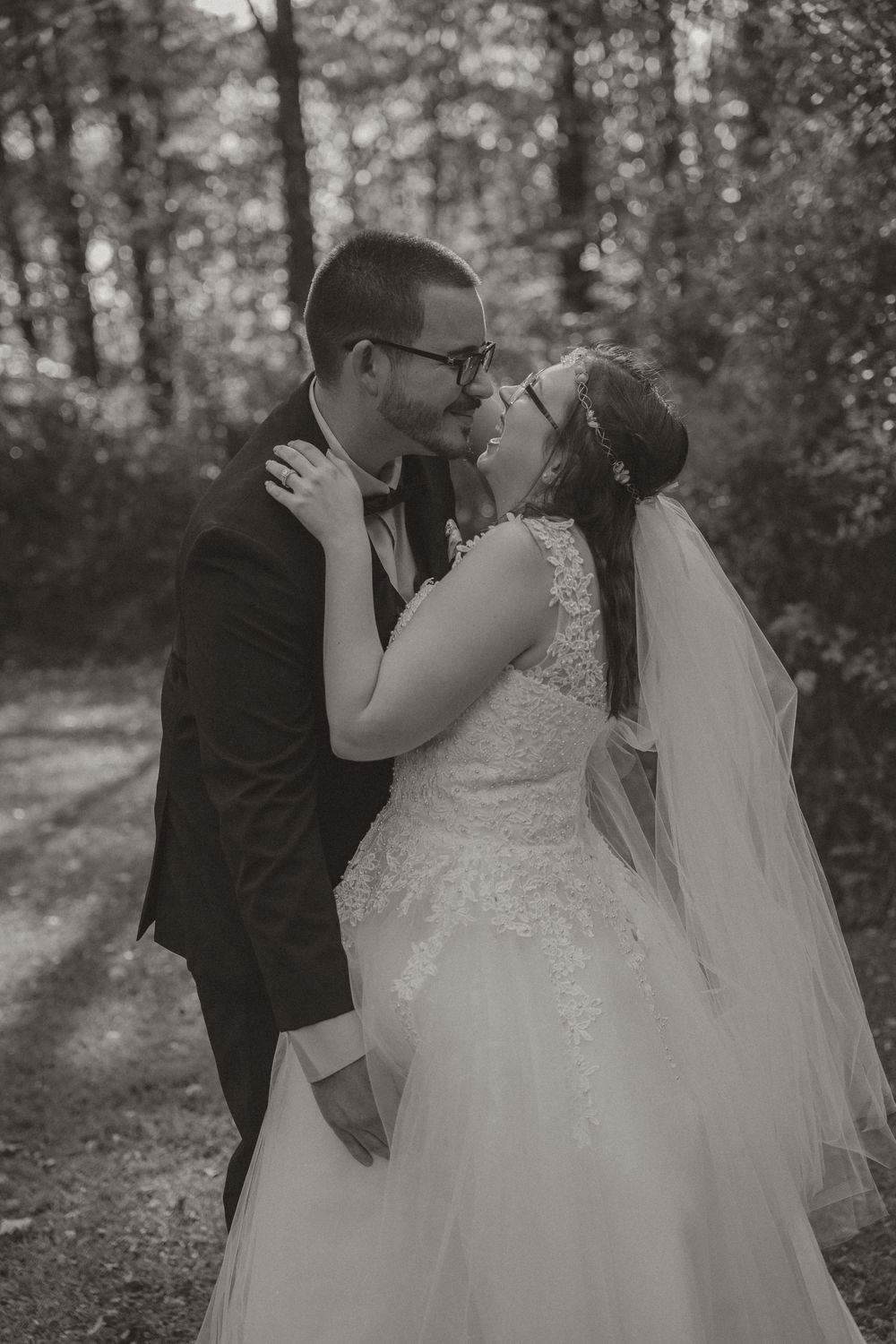 Black and white romantic photo of bride and groom sharing a kiss in a wooded outdoor setting.