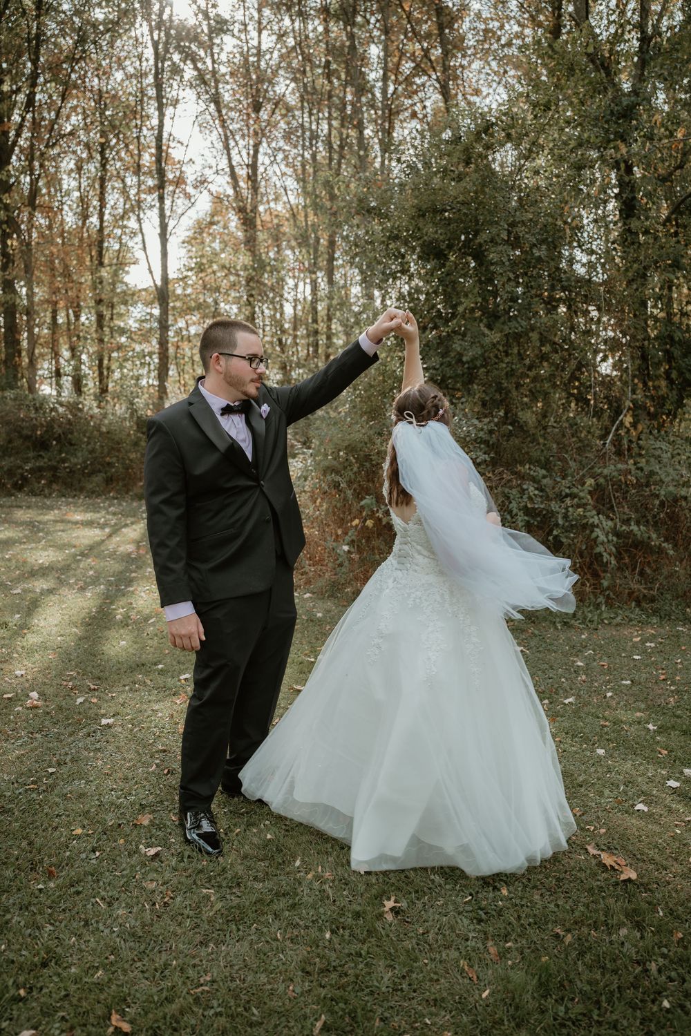 Groom in black tuxedo twirls bride in flowing white gown on a grass lawn with trees in background.