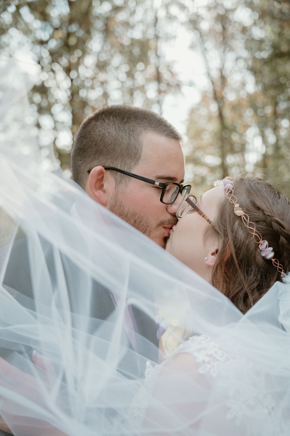 Bride and groom share a romantic kiss beneath a flowing white veil in an outdoor woodland setting.