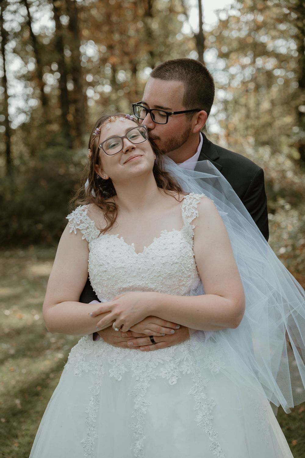 Groom in black suit embraces bride from behind, kissing her cheek as she smiles in a lace wedding gown.