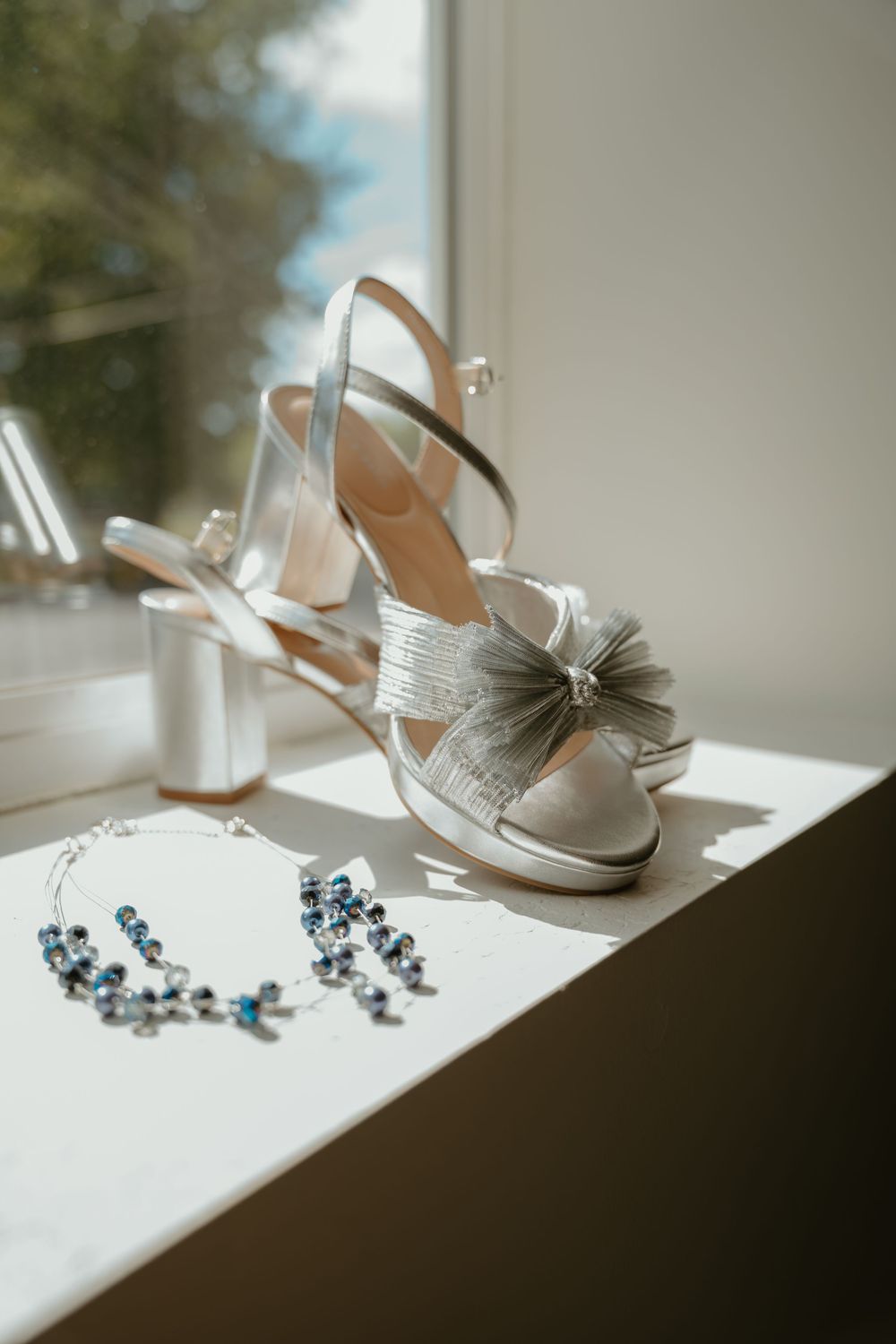 Silver bridal heels with bow detail beside blue crystal jewelry on white windowsill in sunlight.