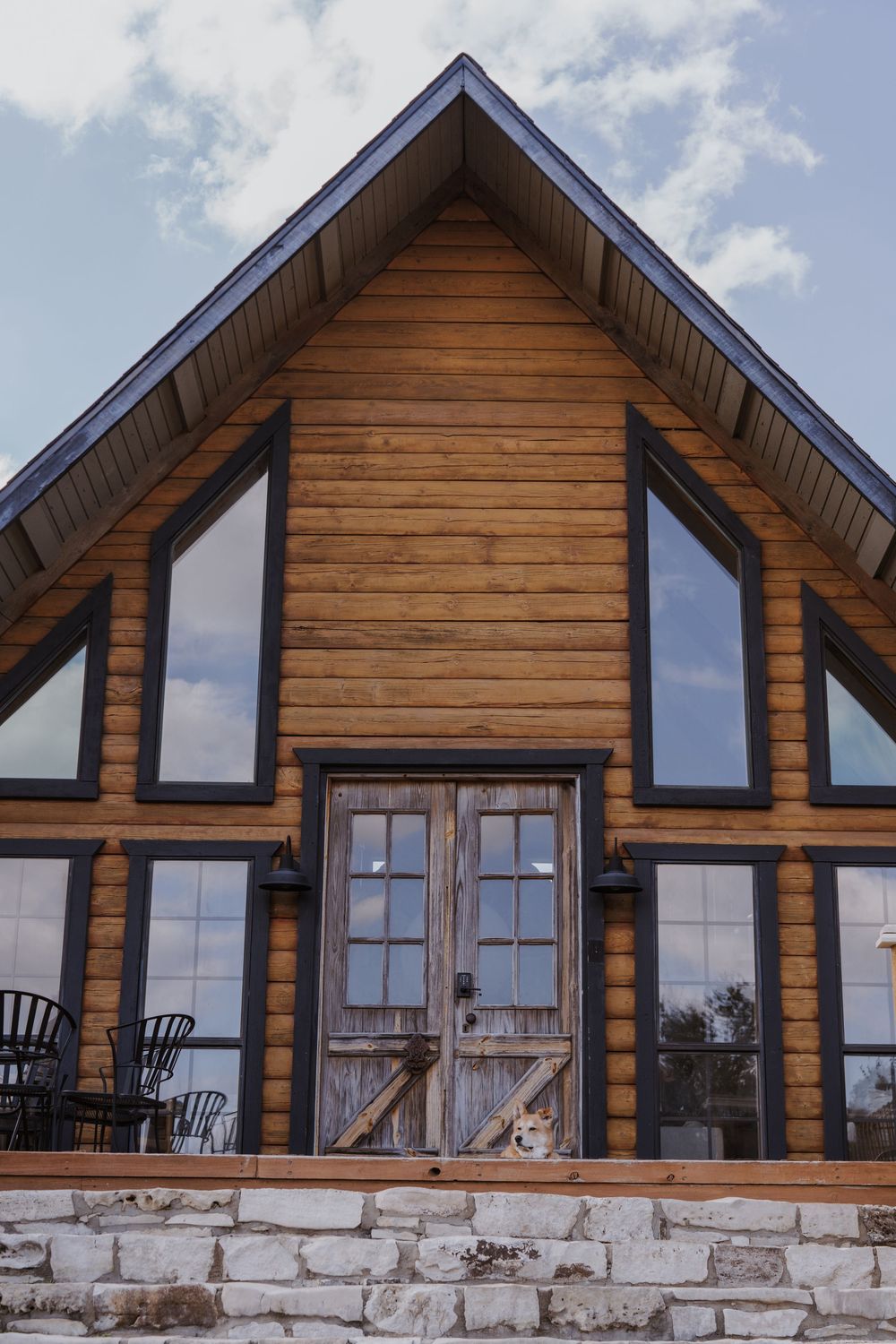 Exterior view of modern log cabin with stone foundation and large windows against blue sky.