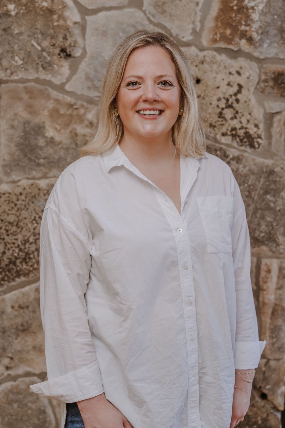 Portrait of a person in a white button-down shirt standing against a stone wall background.