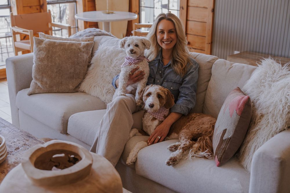 A person sits on a plush couch with two adorable dogs in a rustic cabin setting.