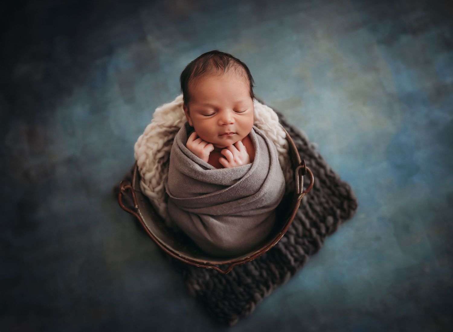A sleeping newborn wrapped in soft gray fabric nestles peacefully in a woven basket against a dreamy blue backdrop.