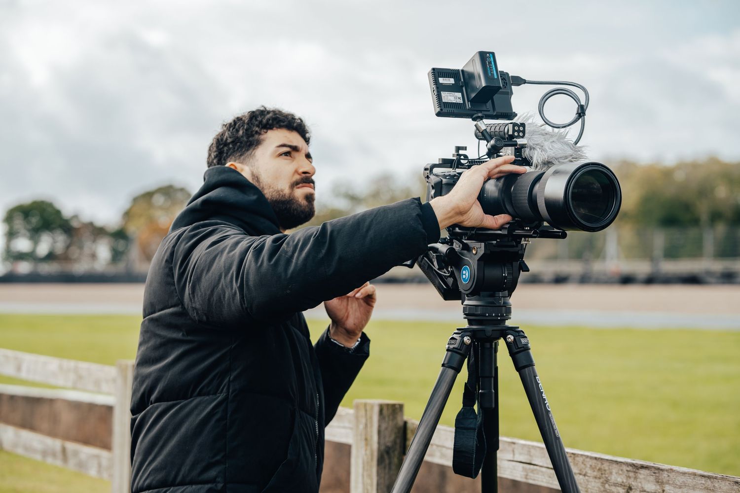 Camera operator films with professional equipment on tripod in outdoor field setting.