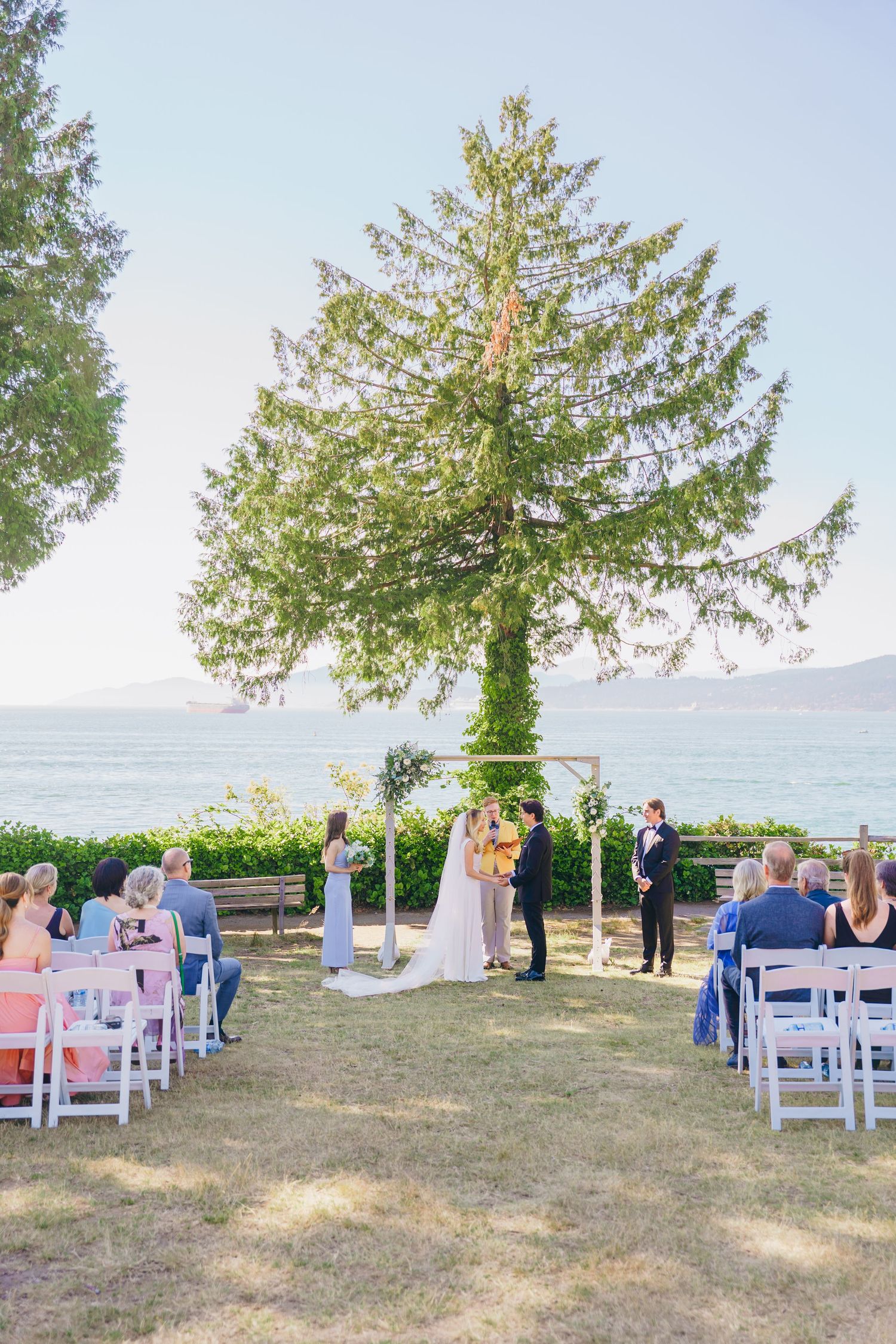 Outdoor wedding ceremony setup on a grassy hillside overlooking the ocean with white chairs and evergreen trees in the background.