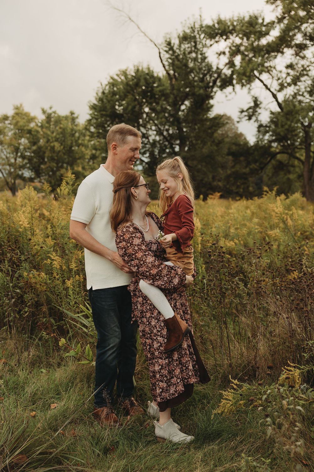 A family embraces together in a sunlit meadow with tall grass and trees during golden hour.