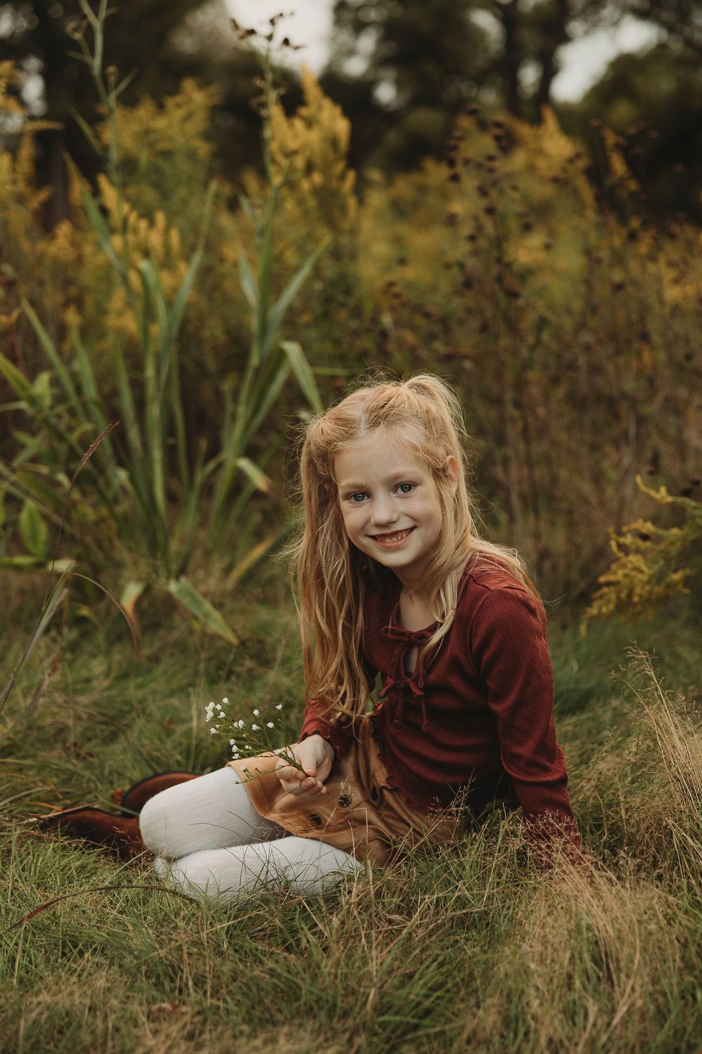 A child in a burgundy sweater lies in autumn grass surrounded by golden foliage during an outdoor photo session.