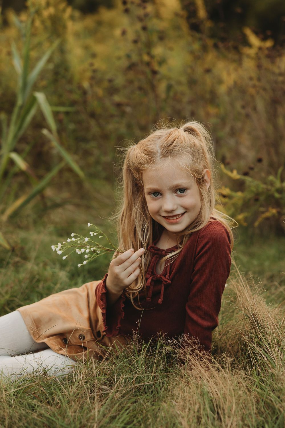 A child in a burgundy sweater lies in autumn grass during an outdoor photo session.