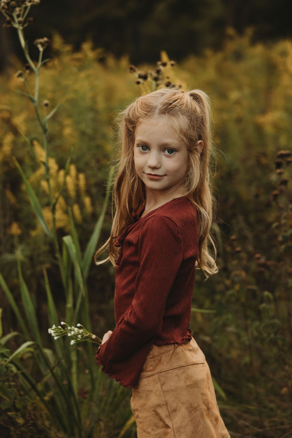 A young child in a burgundy sweater and tan skirt poses playfully in an autumn garden setting.