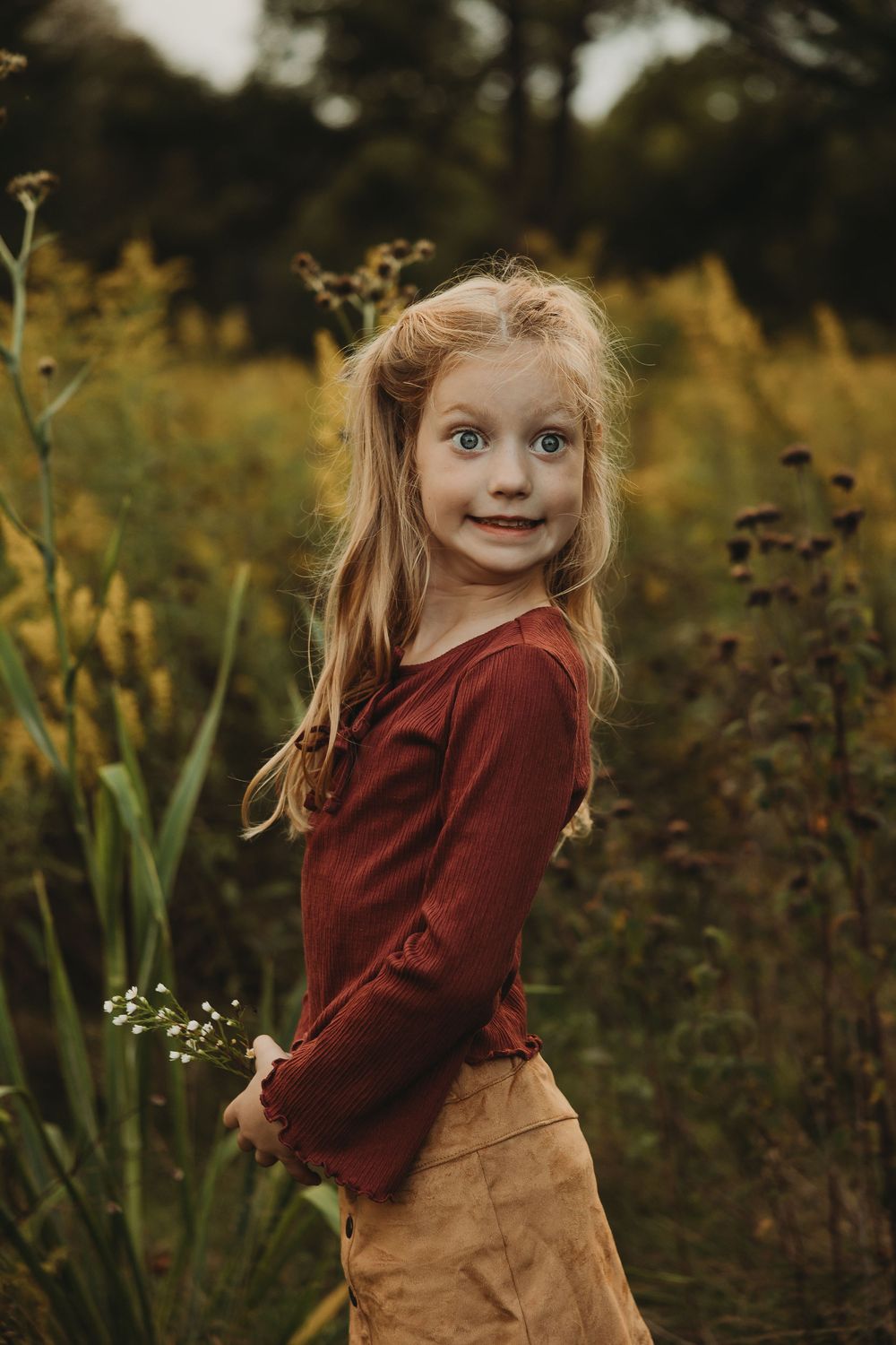 A child in a burgundy sweater and tan skirt twirls playfully in a garden setting during golden hour.