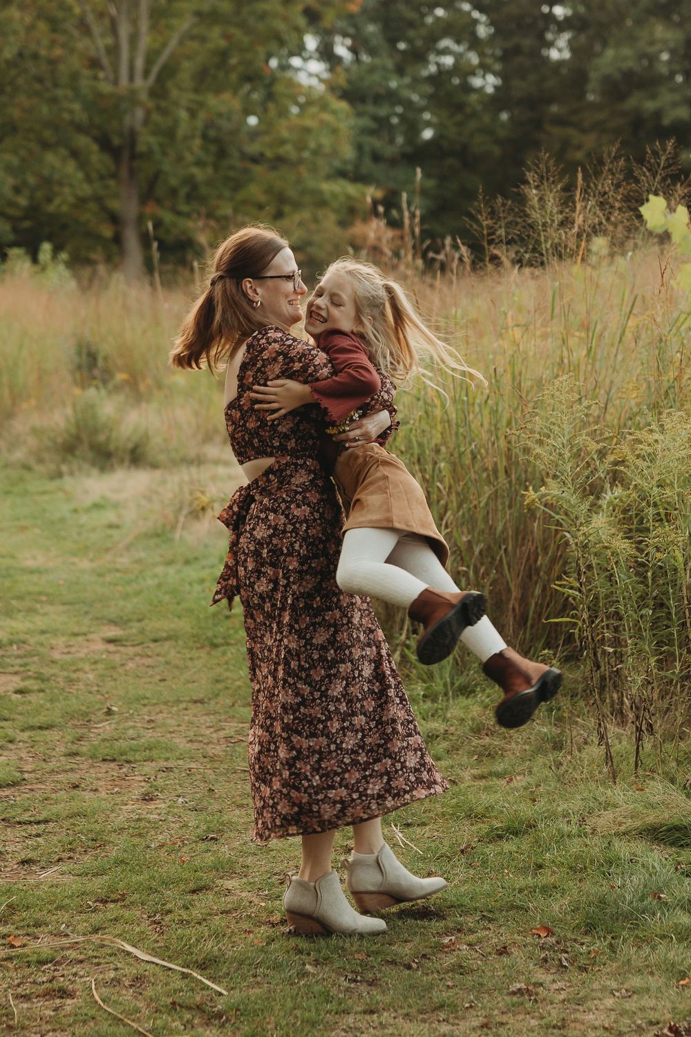 Two people share a joyful embrace in a fall meadow, wearing floral dress and autumn attire against tall grasses.
