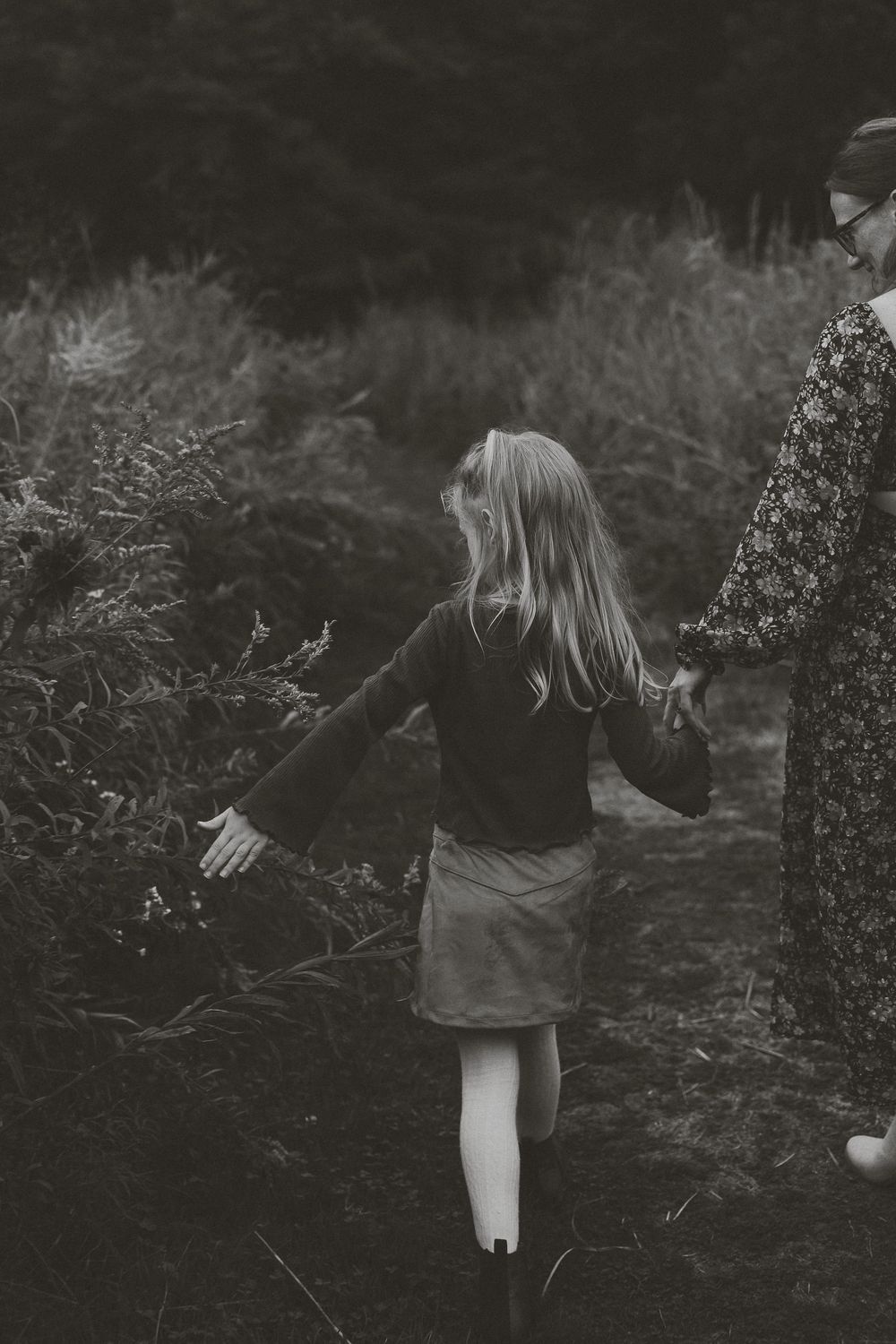 A child in a dark dress walks through a shadowy garden path in a moody black and white photograph.