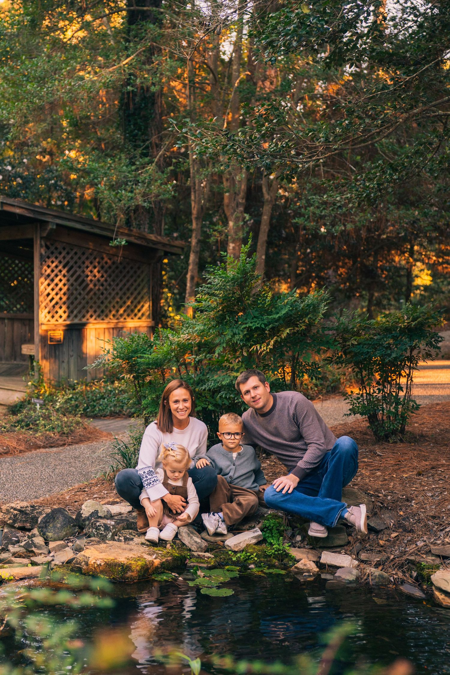 Family sitting together by a garden pond in a wooded backyard during golden hour lighting.