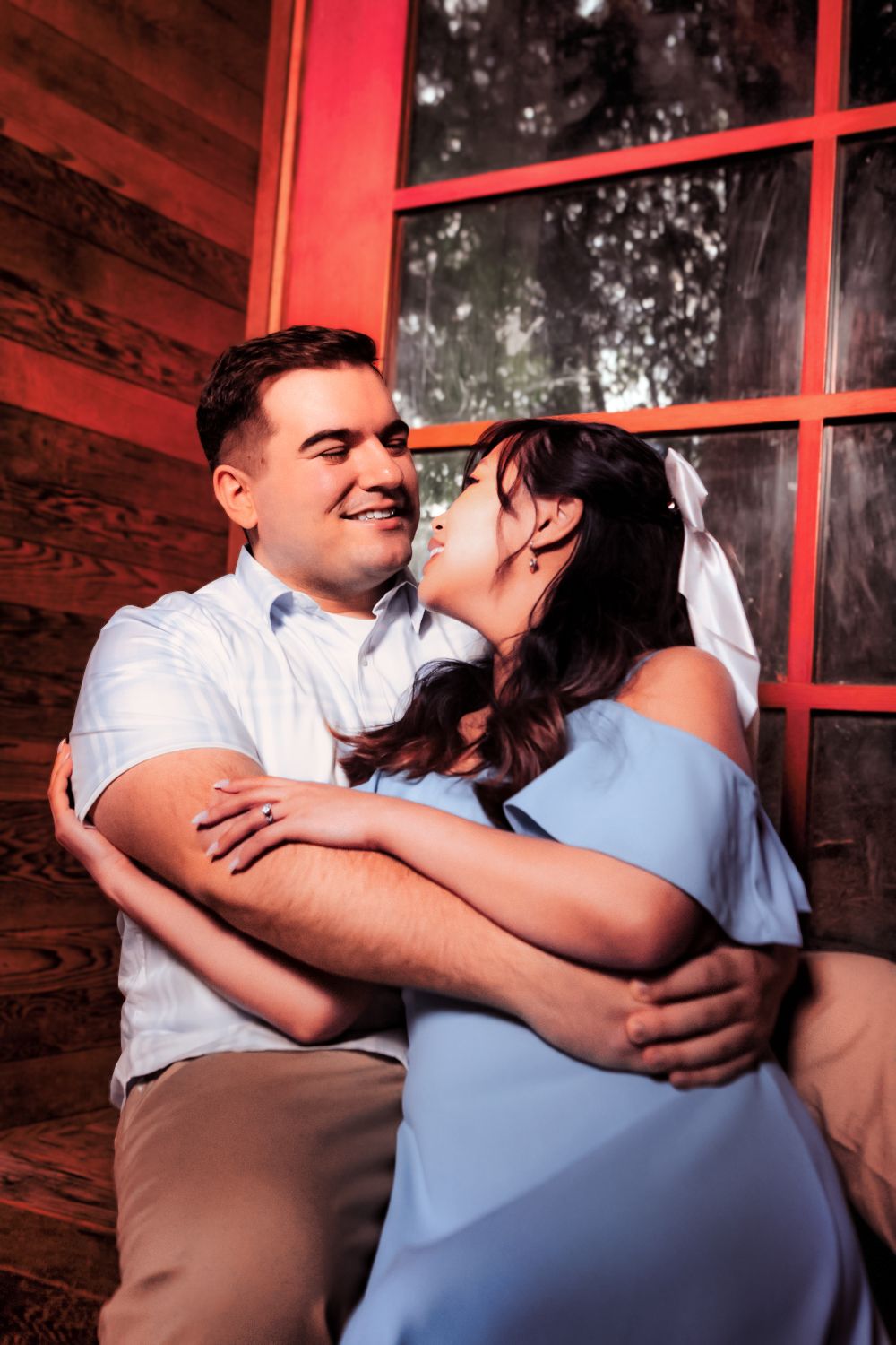 Intimate couple embracing in front of red-framed windows with warm lighting.