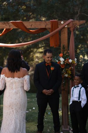 A wedding ceremony takes place under a wooden arbor decorated with flowers in an outdoor garden setting.