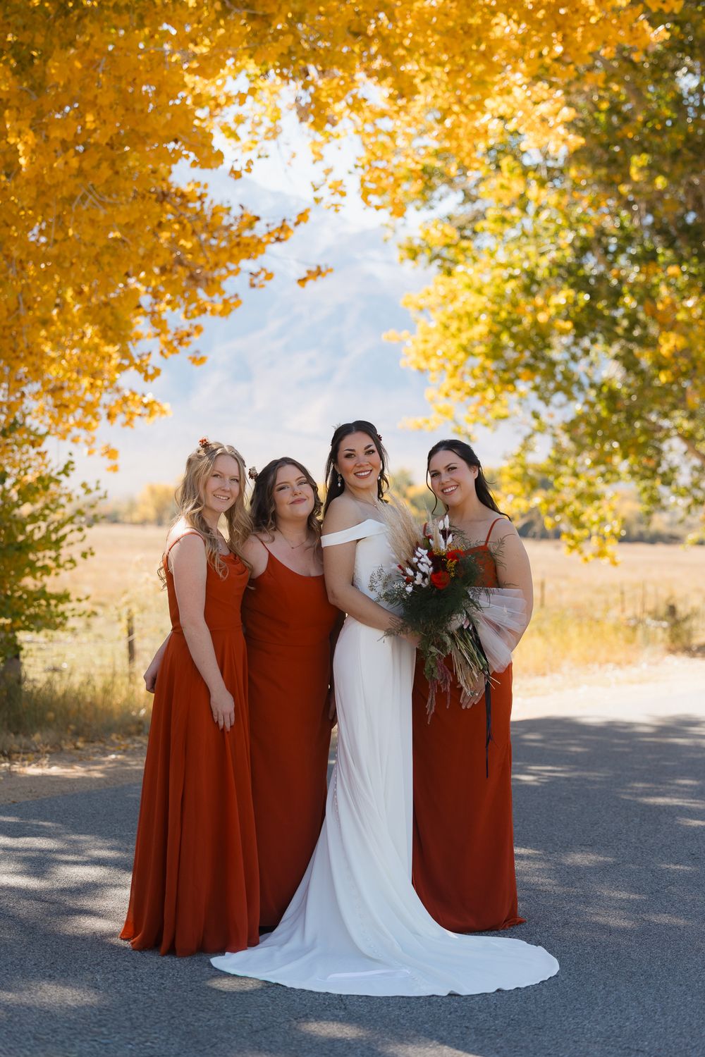 Bride in white gown and bridesmaids in rust-colored dresses pose together under golden autumn trees.