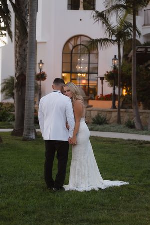 A couple in wedding attire walks together on a grassy lawn near a Spanish-style building at dusk.