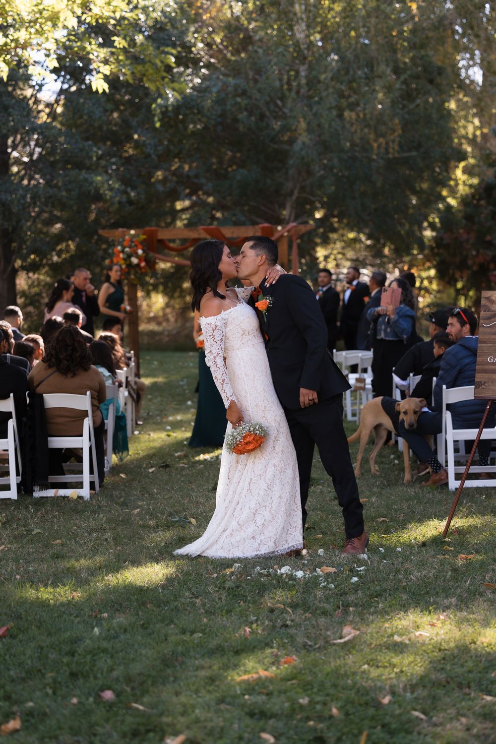 An outdoor wedding ceremony takes place in a garden setting with white chairs and guests gathered under trees at sunset.