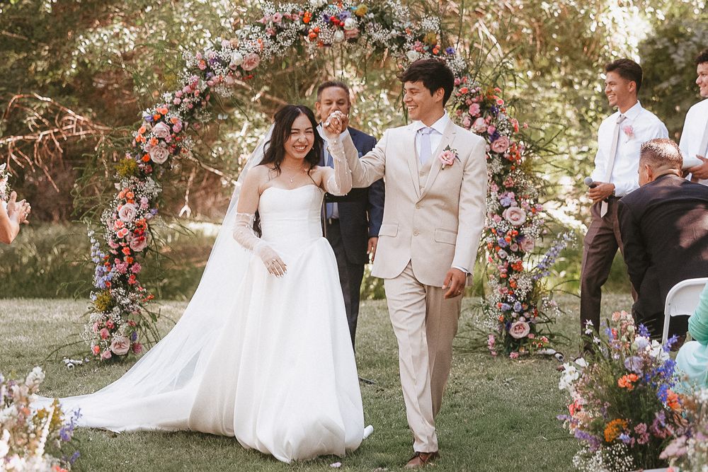 Newlyweds walk under a floral arch during garden wedding ceremony.