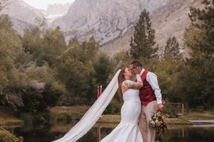 Bride and groom share romantic moment by mountain lake with dramatic backdrop.