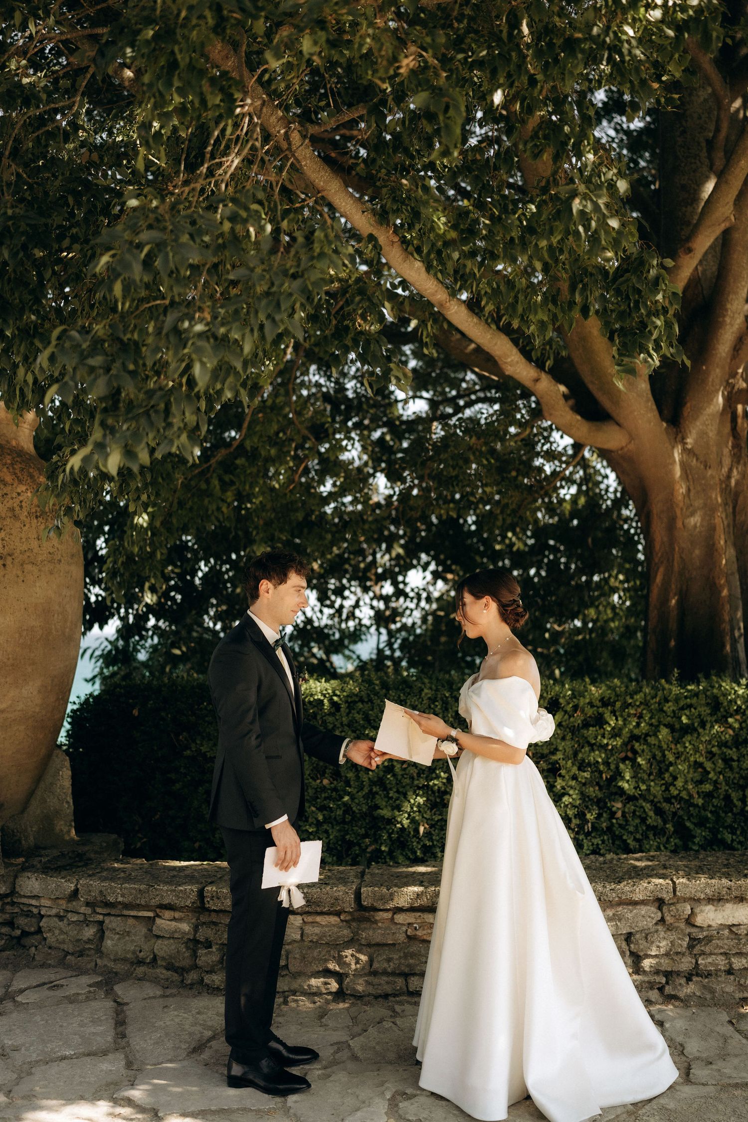 Outdoor wedding ceremony with couple standing beneath trees by a stone wall.