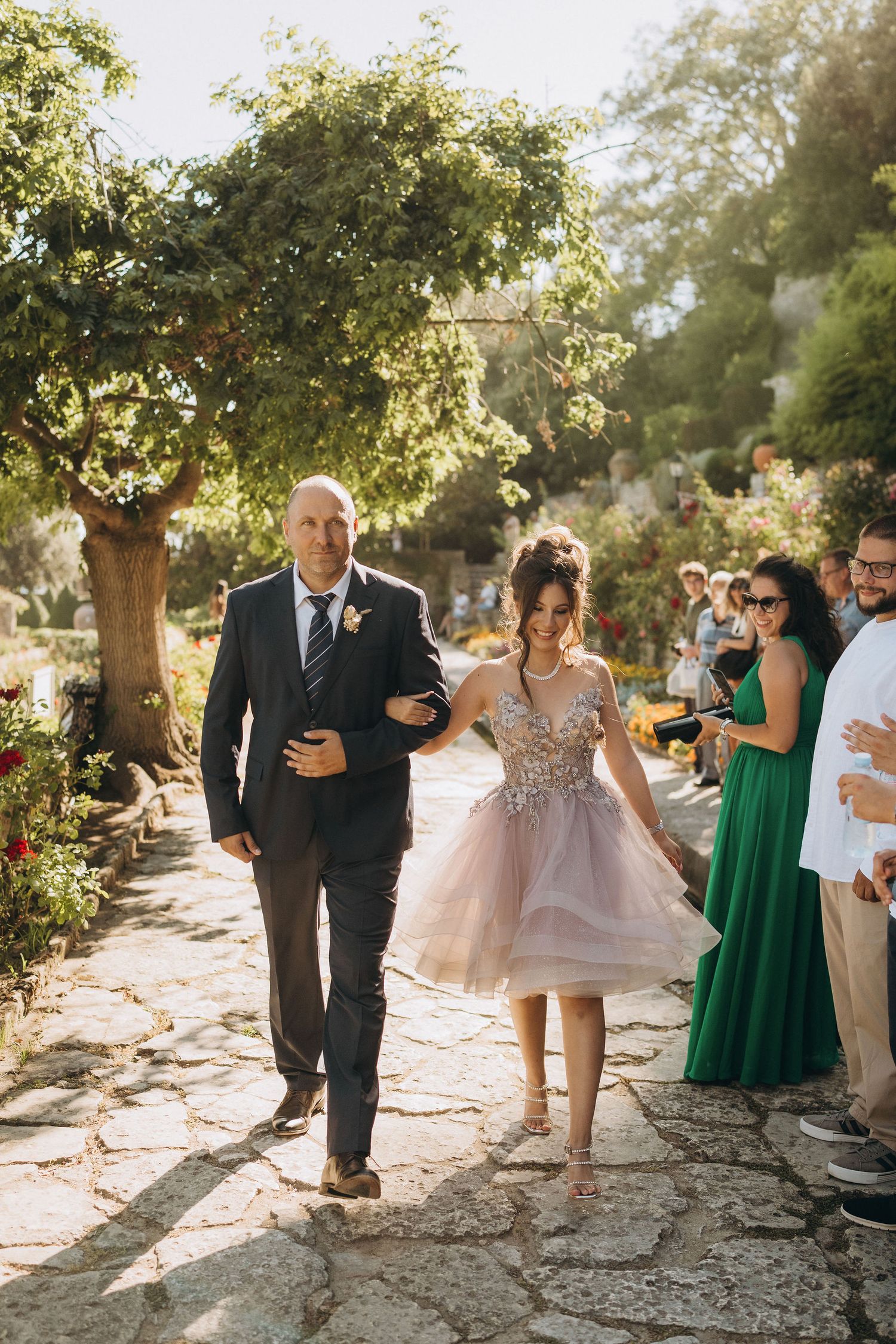 Couple walking down a stone pathway during a wedding ceremony with guests looking on.