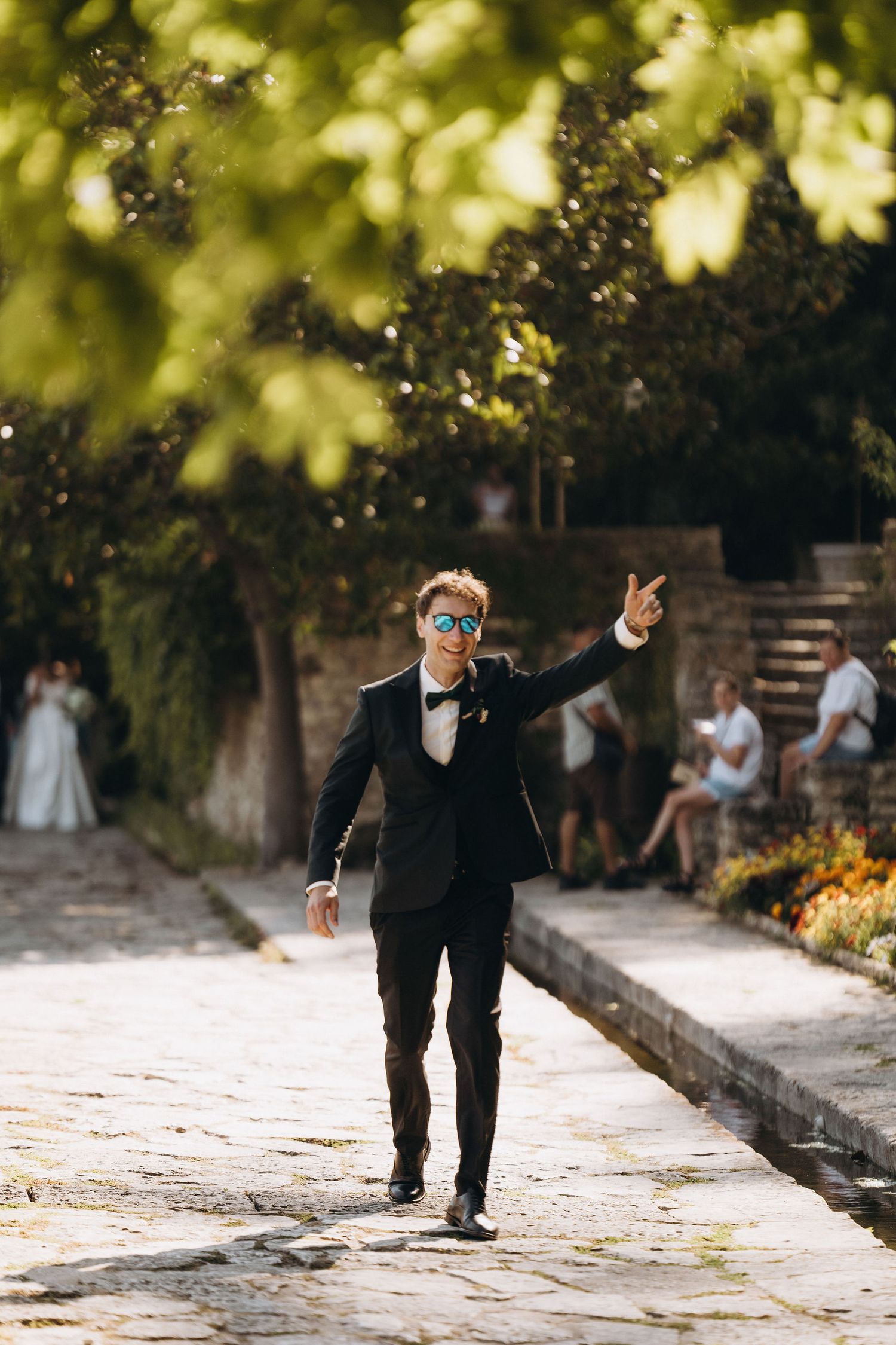 Person in formal attire gesturing while walking along a stone path surrounded by greenery.