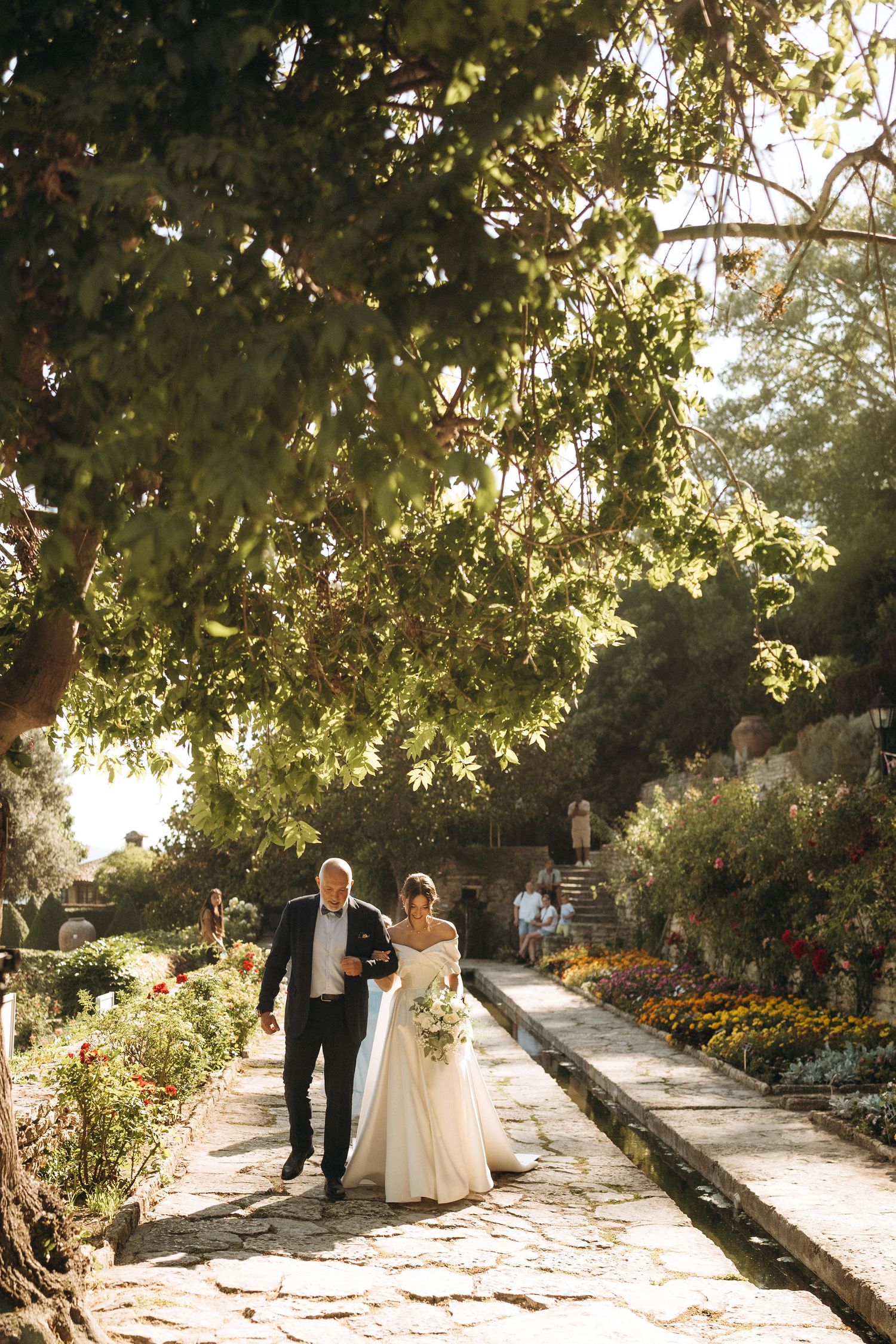 Couple walking down a sunlit garden path with overhanging trees and flowers.