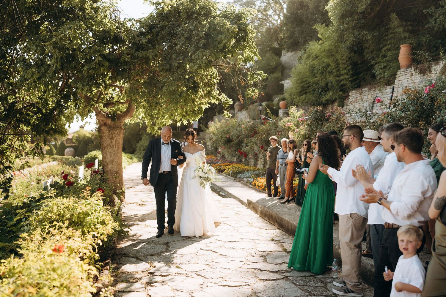 Wedding ceremony procession on a stone pathway lined with guests and lush gardens.