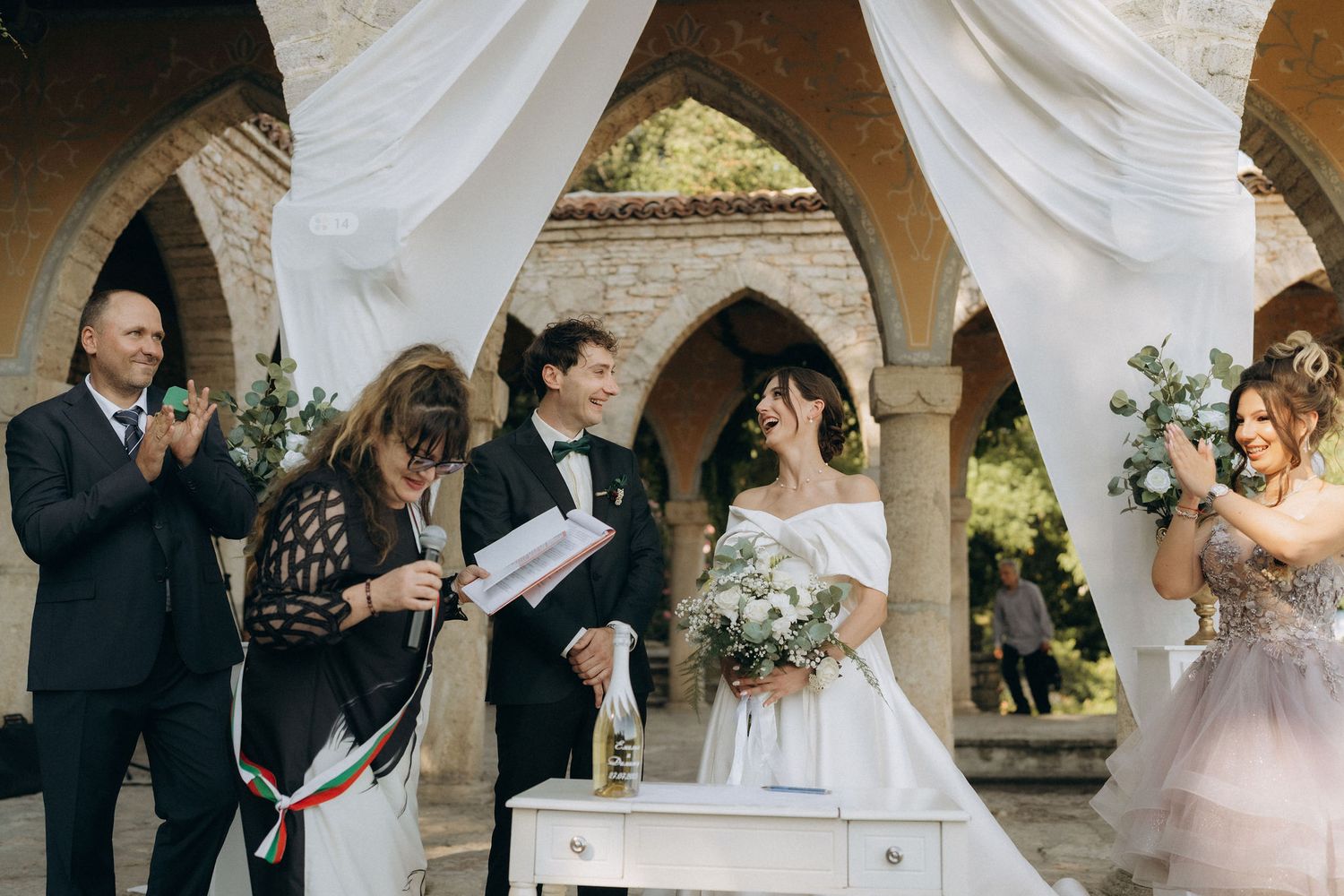 Wedding celebration under stone arches with white fabric decorations and natural lighting.