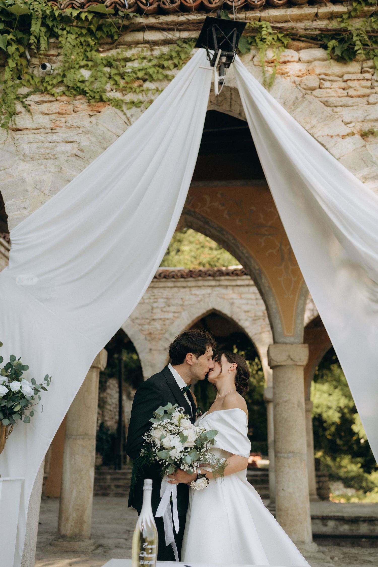 Romantic wedding ceremony moment under draped white fabric and stone archways.