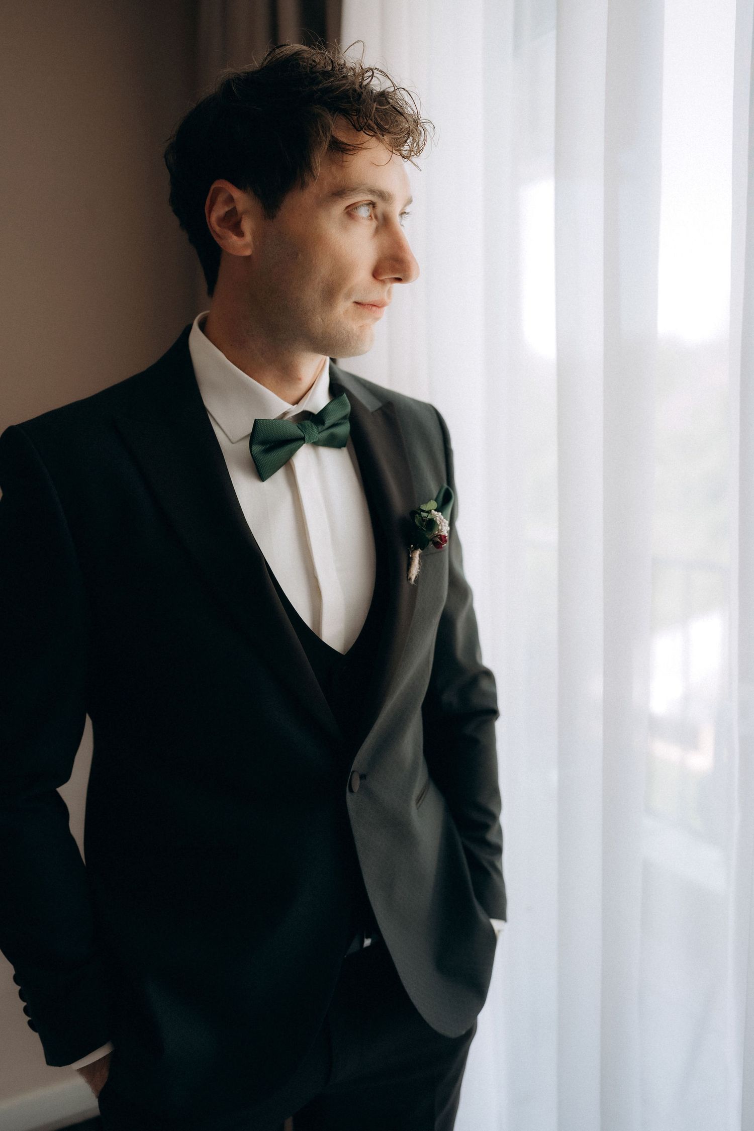 Classic groom portraits in black tuxedo against white window light.
